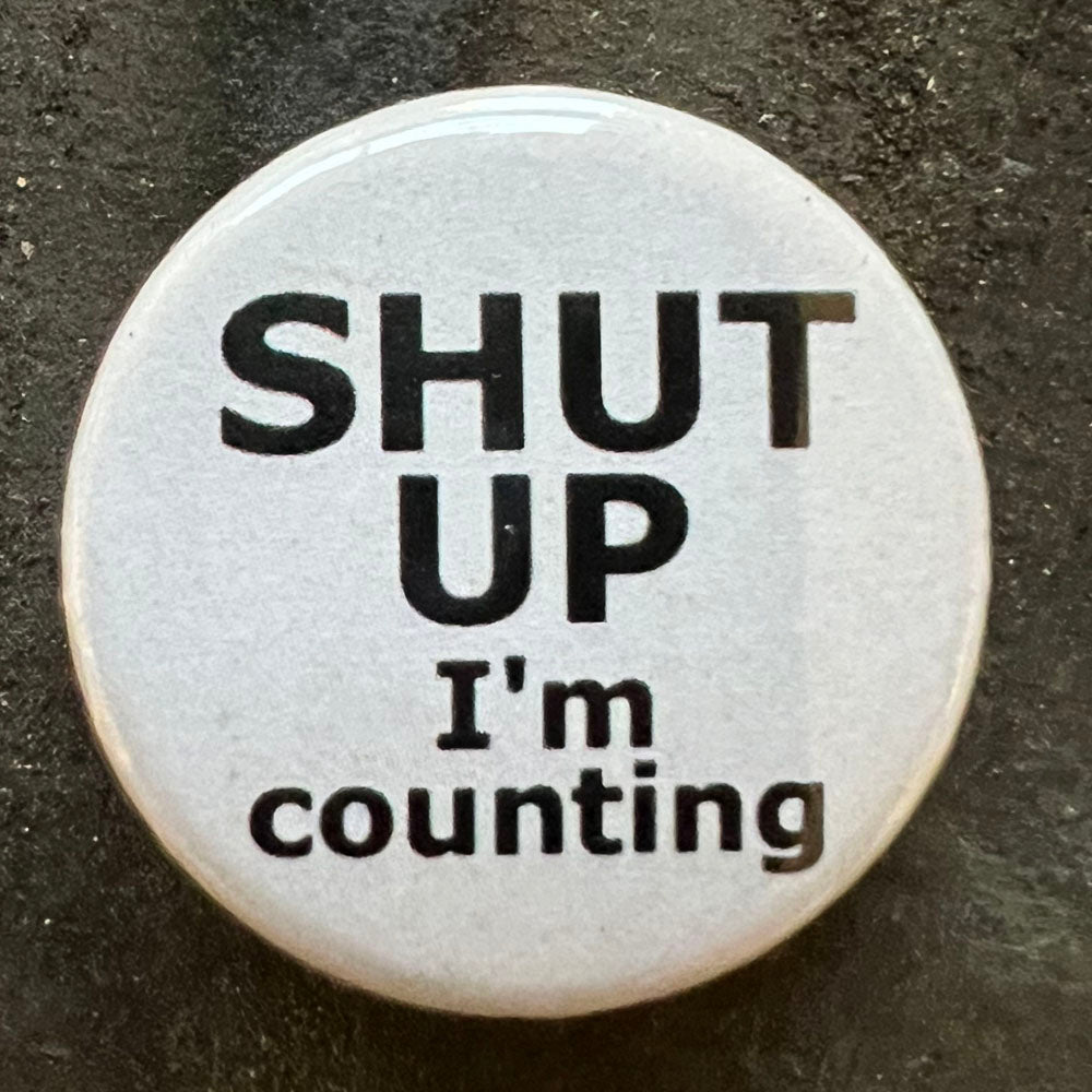 Close-up of a white SHUT UP I'm counting pin badge with black text.
