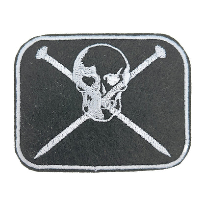 Close-up view of an embroidered felt patch depicting a skull with crossed knitting pins beneath it. The patch features metallic-grey embroidery on a black background, with the skull and knitting pins detailed in metallic-grey thread