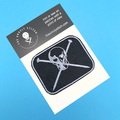 An embroidered felt patch showing a skull with crossed knitting pins beneath it, presented on a card. The patch has silver-grey embroidery on a black background, with the skull and knitting pins detailed in silver-grey thread. The card has 'The Unruly Stitch' branding with instructions to iron or sew on the patch.