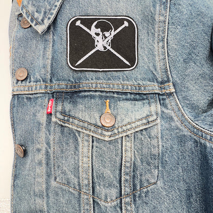 Patch on jacket: An embroidered felt patch featuring a skull with crossed knitting pins beneath it, attached to the pocket area of a denim jacket. The patch has silver-grey embroidery on a black background, with the skull and knitting pins detailed in silver-grey thread, adding a unique accent to the jacket.