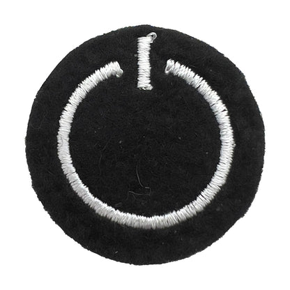 Close-up of a felt badge with an embroidered power button symbol in white threads on a black background, designed by The Unruly Stitch.