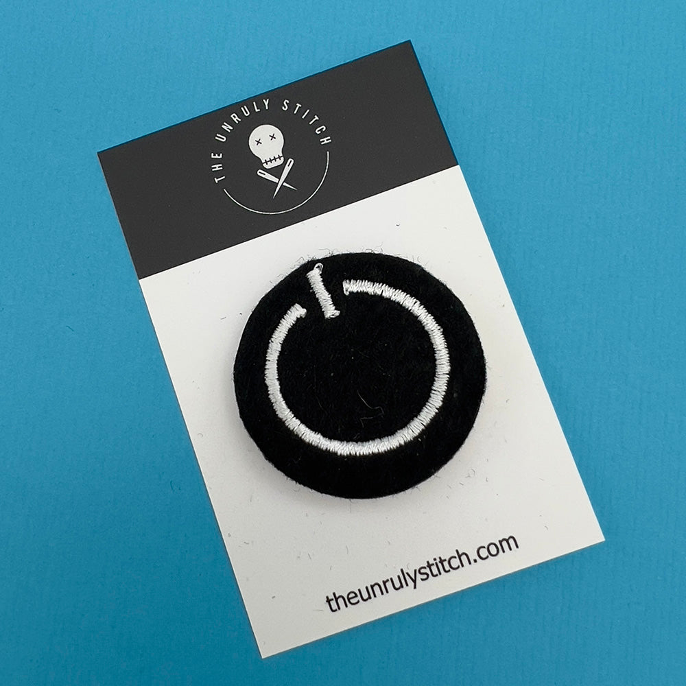 Embroidered felt badge depicting a power button symbol in white threads on a black background, mounted on a branded card from The Unruly Stitch