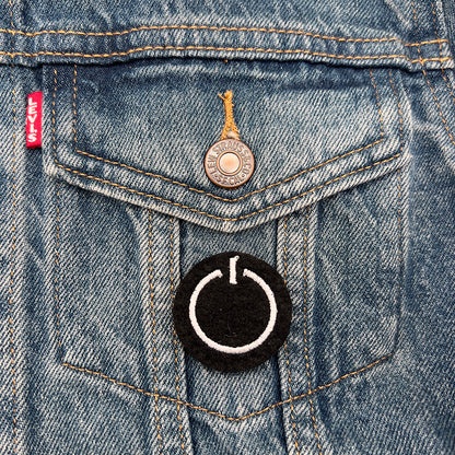 Embroidered felt badge with a power button symbol design in white threads on a black background, pinned on the pocket of a denim jacket, designed by The Unruly Stitch.
