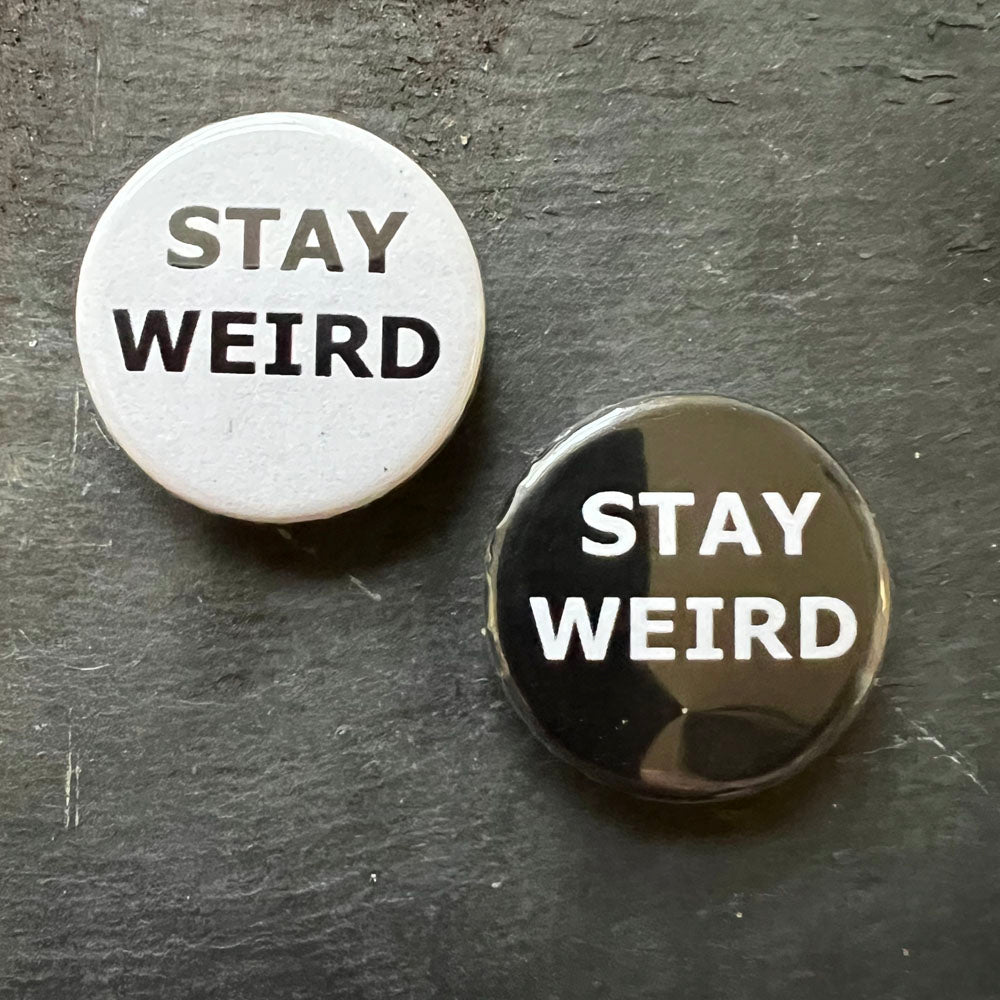 Two "Stay Weird" pin badges, one in white and one in black, with white text on a dark background.
