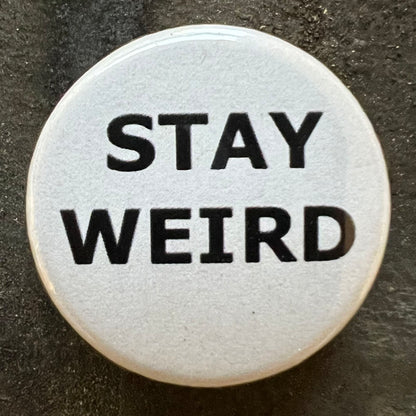 Close-up of a white "Stay Weird" pin badge with black text on a dark background.