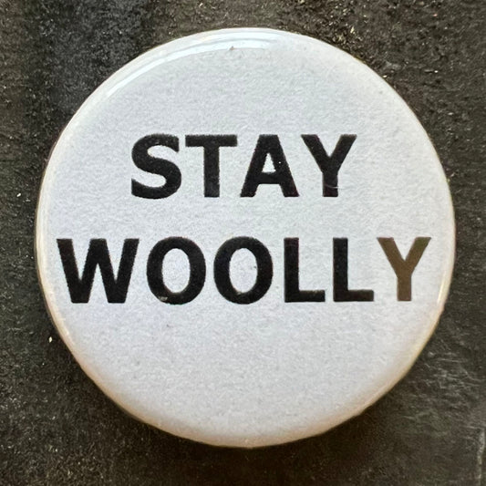 Close-up of a white STAY WOOLLY pin badge with black text.