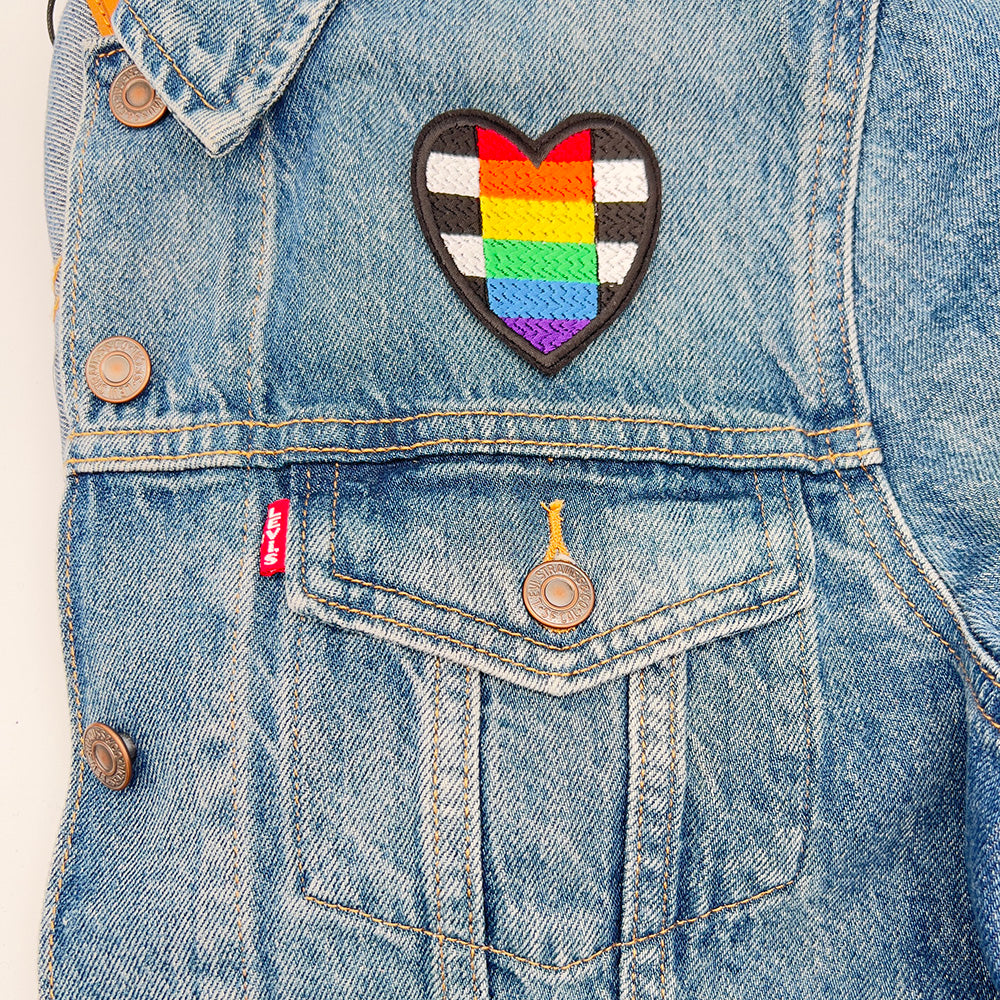 Heart-shaped straight allies pride flag patch ironed onto a denim jacket above the left-side pocket.