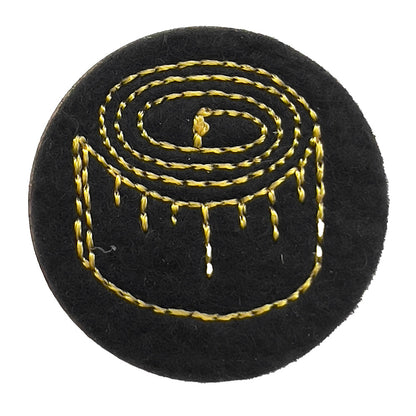 Close-up of a felt badge with an embroidered tape measure in yellow threads on a black background, designed by The Unruly Stitch.