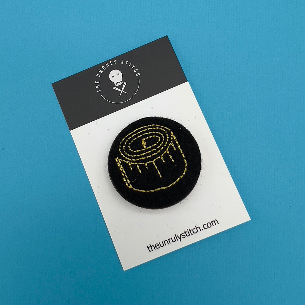 Embroidered felt badge depicting a tape measure in yellow threads on a black background, mounted on a branded card from The Unruly Stitch.