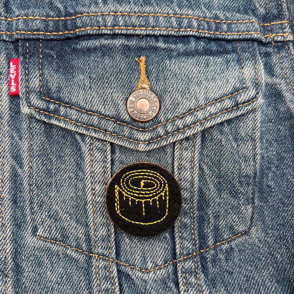 Embroidered felt badge with a tape measure design in yellow threads on a black background, pinned on the pocket of a denim jacket, designed by The Unruly Stitch.