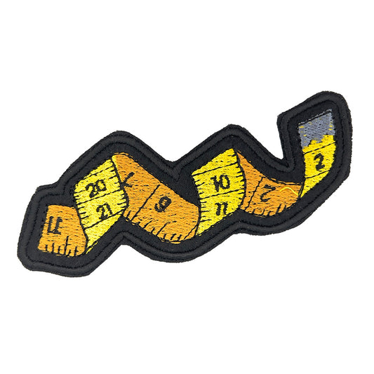 Close-up image: Close-up view of an embroidered felt patch shaped like a winding tape measure. The patch features a tape measure with numbers ranging from 1 to 12, depicted in yellow and orange sections with black outlines and details.