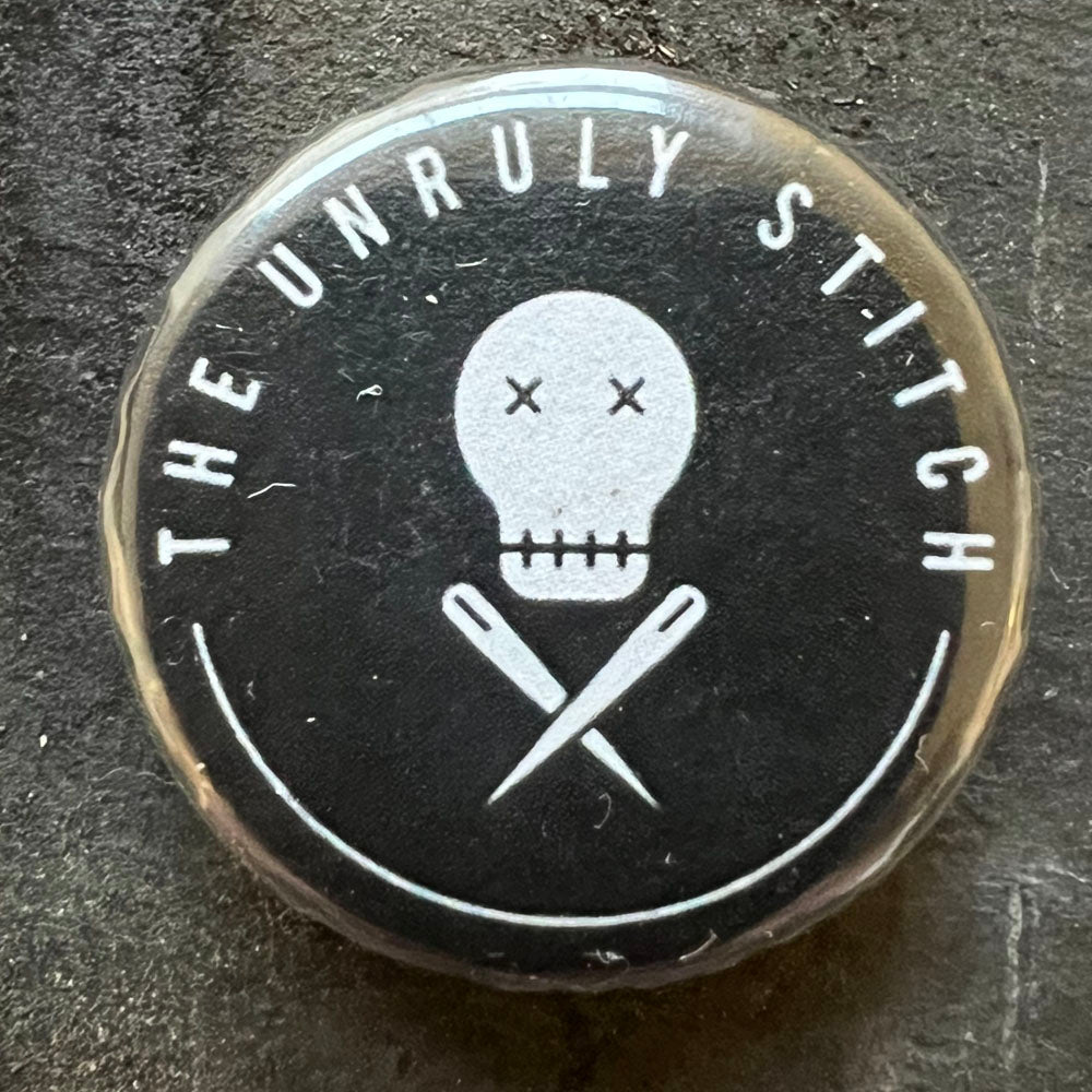 Close-up of a black pin badge with a white skull and crossed knitting needles design, with the text "THE UNRULY STITCH" in white letters around the edge.