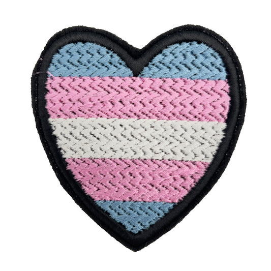 A close-up view of a heart-shaped patch with the Transgender Pride flag colors, featuring light blue, pink, and white horizontal stripes. The patch has a black embroidered border.