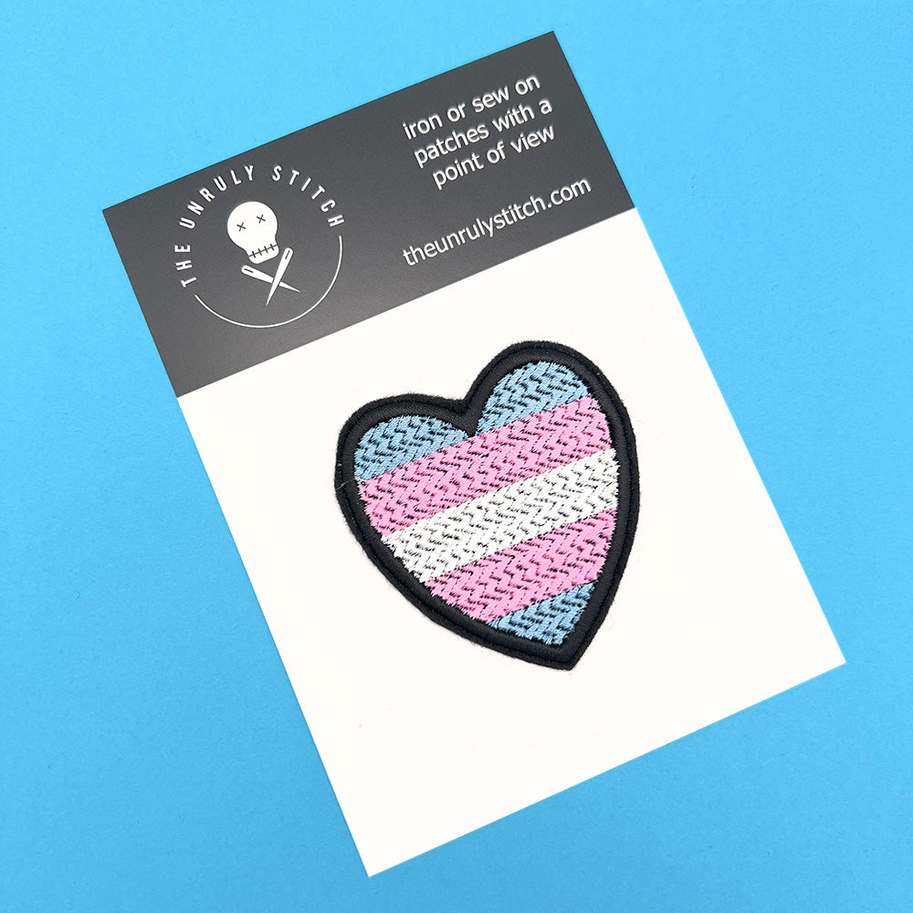  A heart-shaped Transgender Pride flag patch with light blue, pink, and white horizontal stripes, attached to a white card. The card sits on a blue background and includes branding details of "The Unruly Stitch."