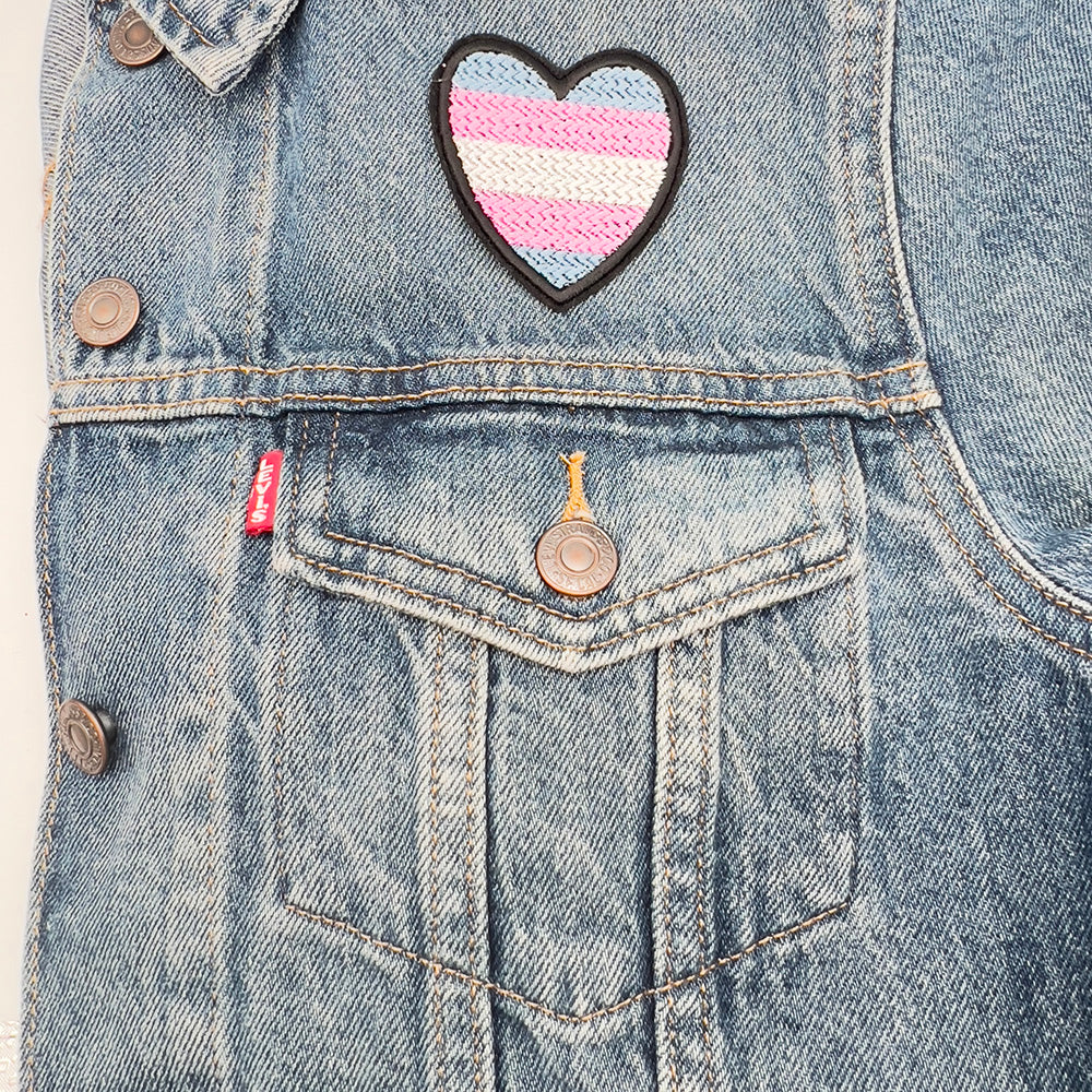 A heart-shaped patch with the Transgender Pride flag colors (light blue, pink, and white horizontal stripes) ironed above the pocket of a denim jacket, displaying the patch prominently on the upper section of the jacket.