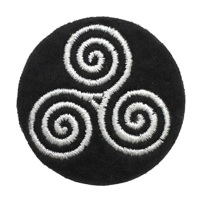 Close-up of a felt badge with an embroidered triskelion infinity symbol in white threads on a black background, designed by The Unruly Stitch
