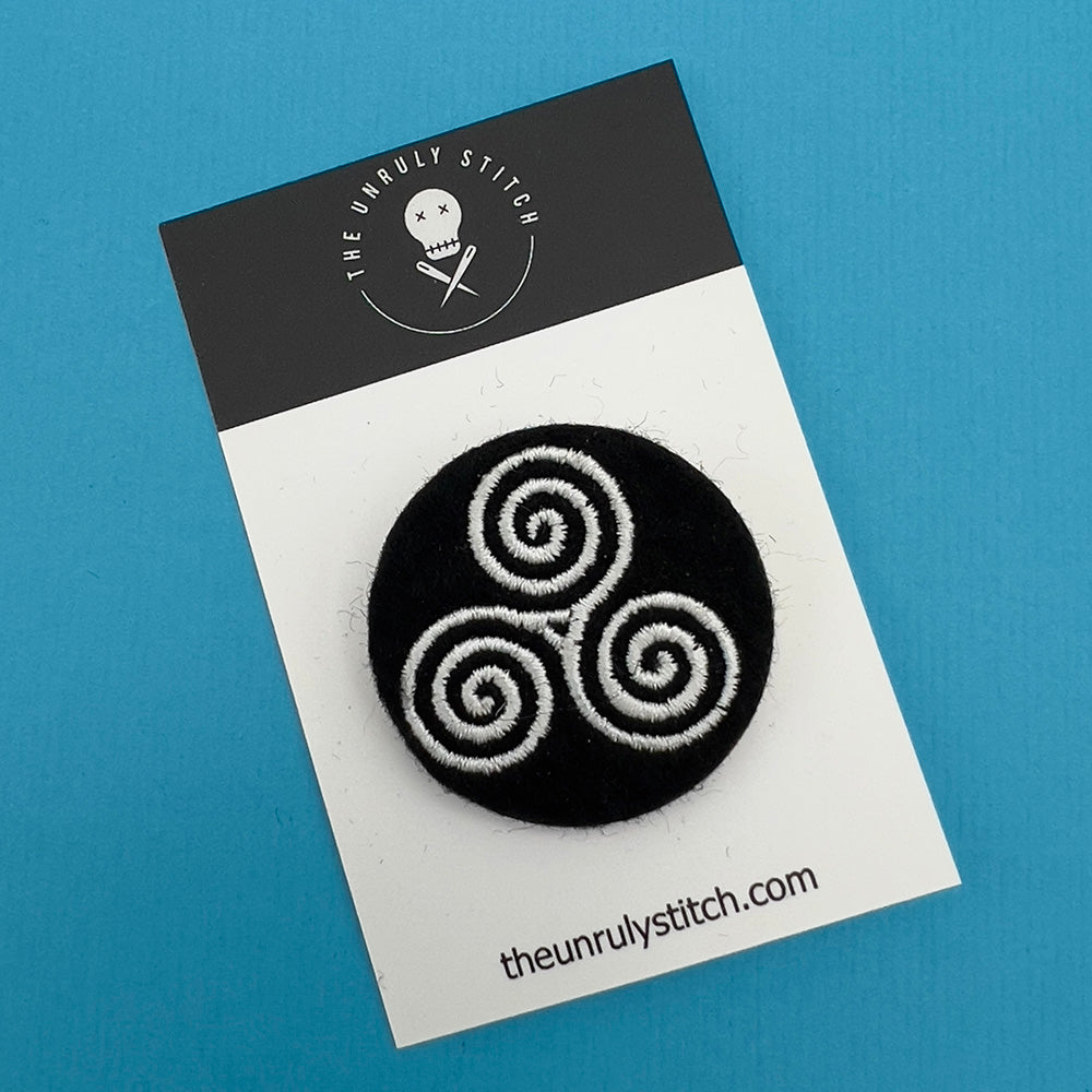 Embroidered felt badge depicting a triskelion infinity symbol in white threads on a black background, mounted on a branded card from The Unruly Stitch.