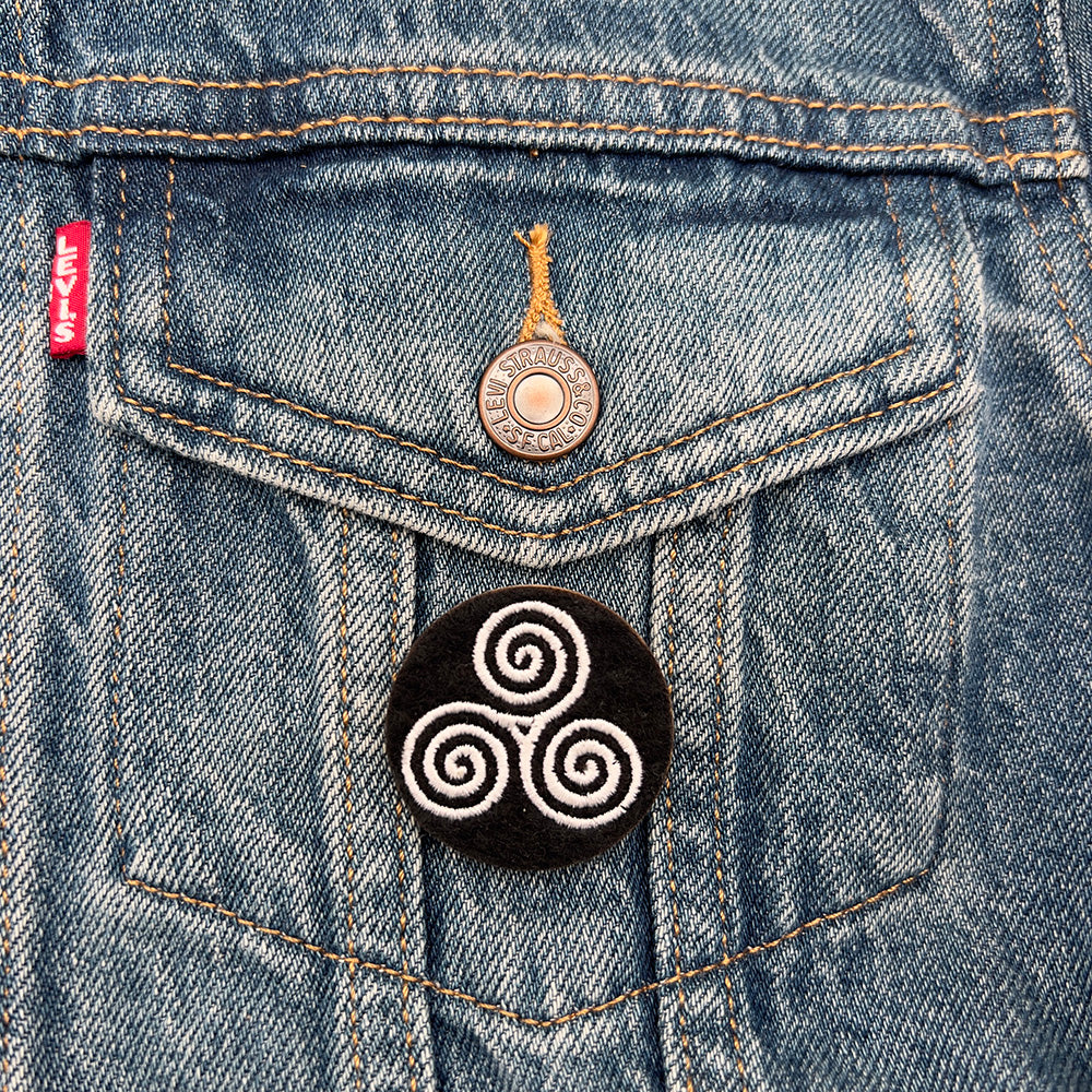 Embroidered felt badge with a triskelion infinity symbol design in white threads on a black background, pinned on the pocket of a denim jacket, designed by The Unruly Stitch.