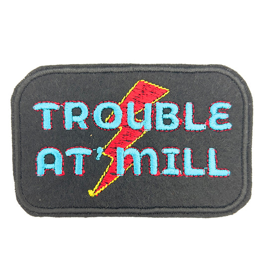 A close-up of an embroidered patch on black felt in the shape of a rectangle with rounded corners. The patch features the text "TROUBLE AT 'MILL" in blue and red letters, with a red and yellow lightning bolt in the background.