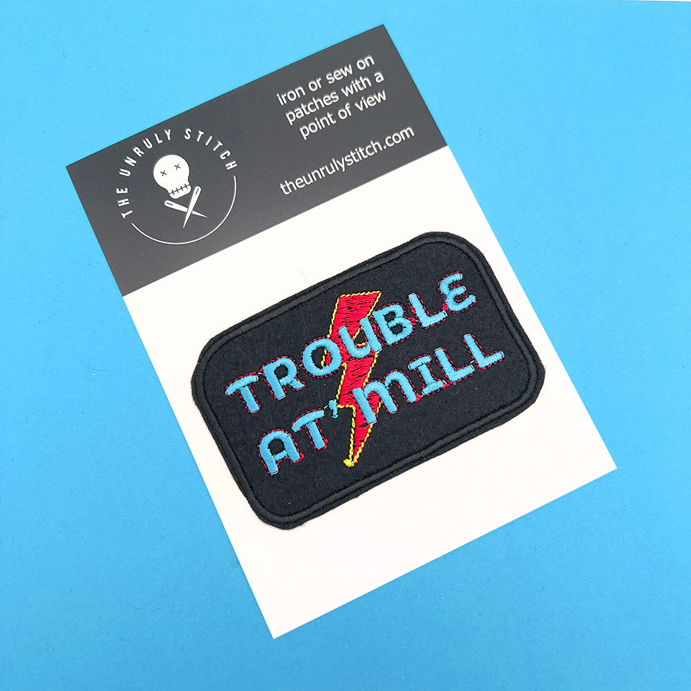 An embroidered patch on a white card with a black header from "The Unruly Stitch." The patch is rectangular with rounded corners, featuring the text "TROUBLE AT 'MILL" in blue and red letters, and a red and yellow lightning bolt in the background. The card sits on a blue background.