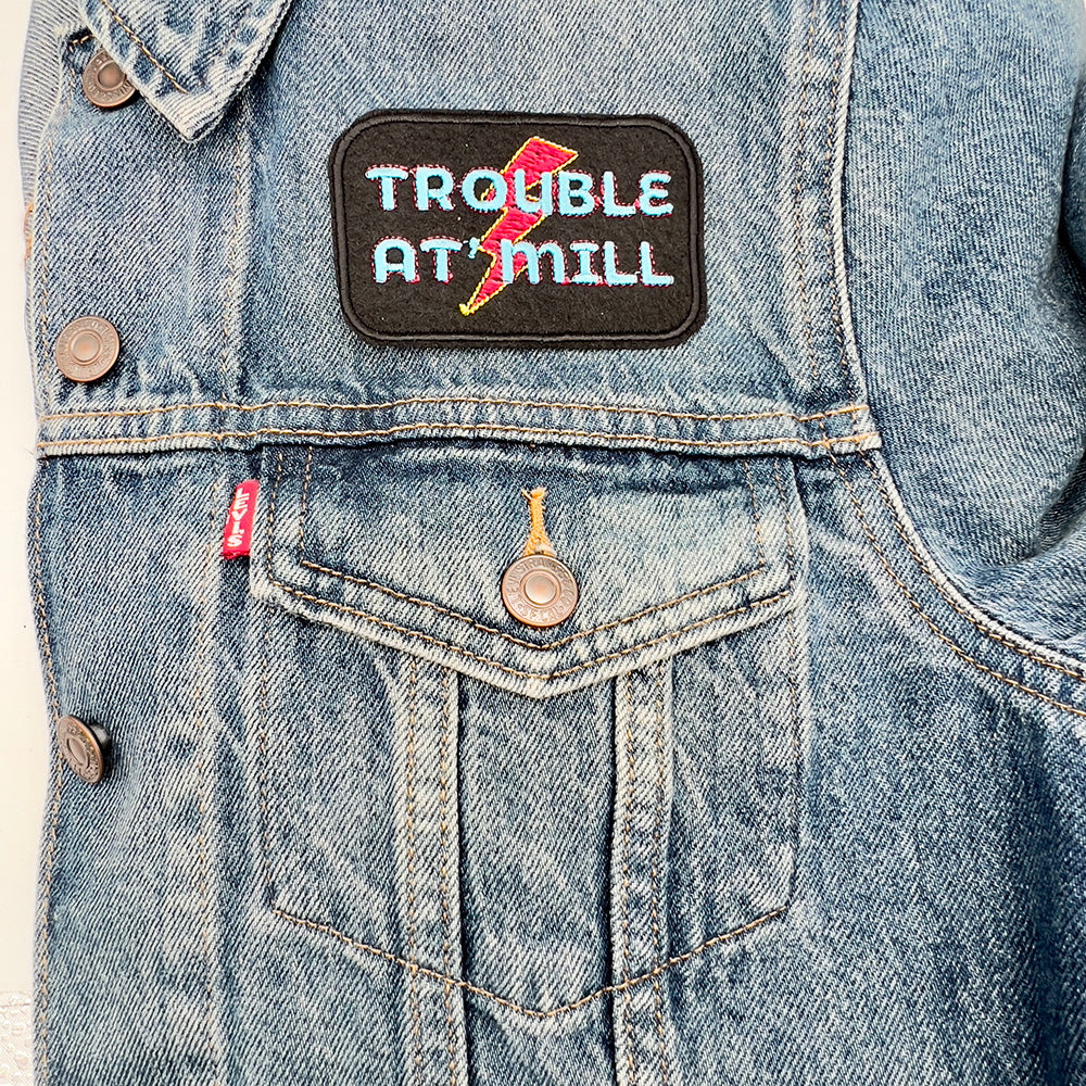 A rectangular embroidered patch with rounded corners featuring the text "TROUBLE AT 'MILL" in blue and red letters, and a red and yellow lightning bolt in the background. The patch is ironed onto a denim jacket above the pocket on the left side.