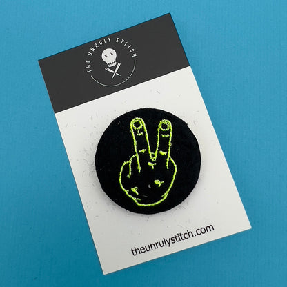 Branded card with felt badge with an embroidered hand with two fingers up in yellow threads on a black background, designed by The Unruly Stitch.