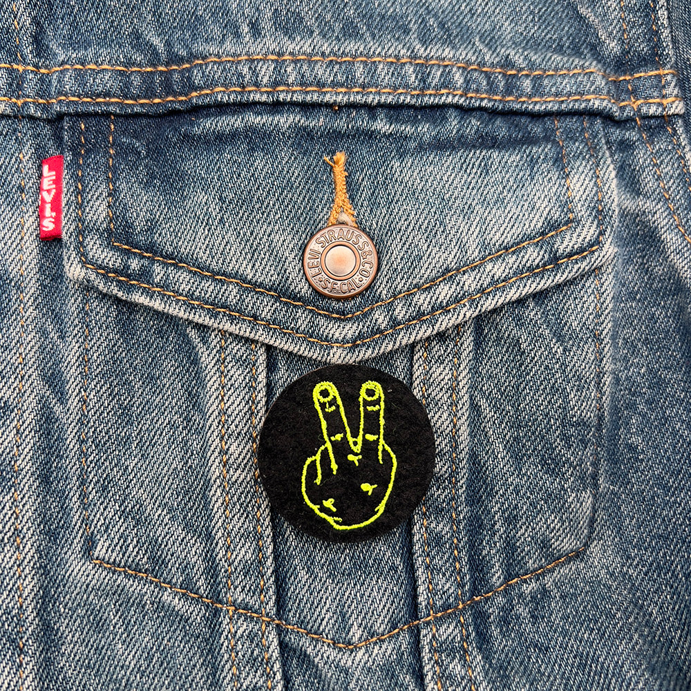 Felt badge with an embroidered hand with two fingers up in yellow threads on a black background, designed by The Unruly Stitch. Badge is pinned to the pocket of a denim jacket.
