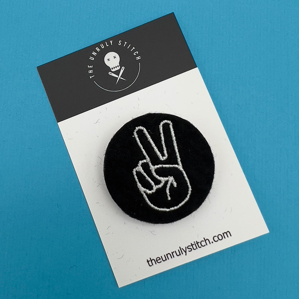 Embroidered felt badge depicting a peace or victory hand sign in white threads on a black background, mounted on a branded card from The Unruly Stitch.