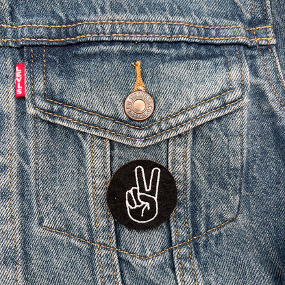 Embroidered felt badge with a peace or victory hand sign design in white threads on a black background, pinned on the pocket of a denim jacket, designed by The Unruly Stitch.