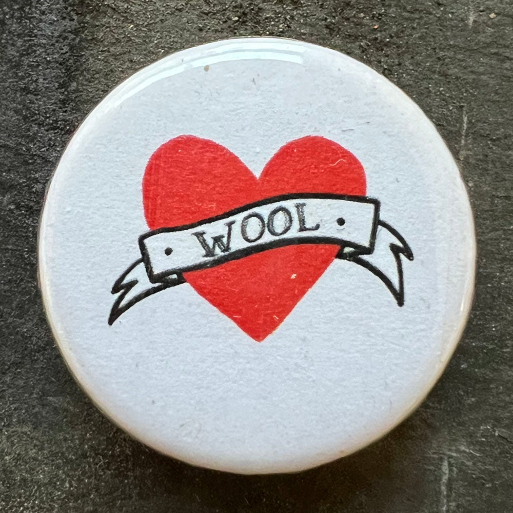 Close-up of a white pin badge with a red heart and the word "WOOL" in a banner across it