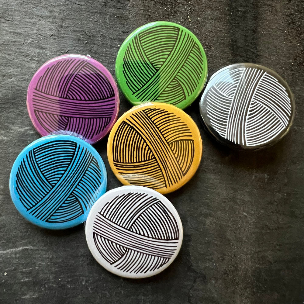 Collection of colorful 'Yarn Sniffer' pin badges in black, blue, green, purple, yellow, and white, displayed on a dark slate background.
