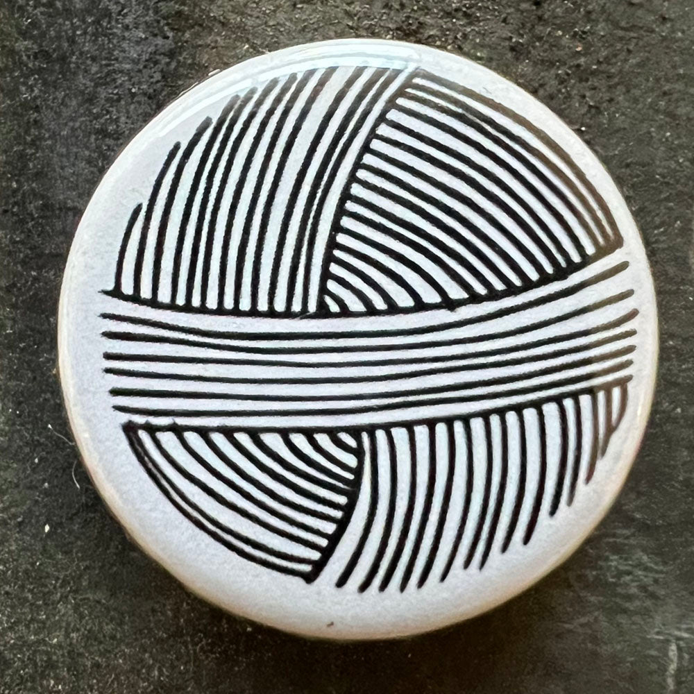 Close-up of a white pin badge featuring a black yarn ball design, displayed on a dark slate background