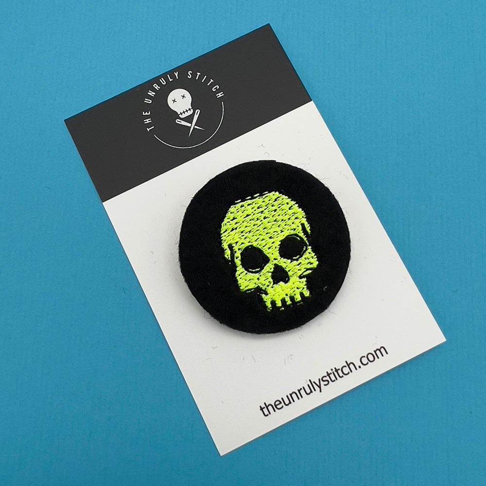 A black felt badge with a yellow embroidered skull displayed on a branded card. The card has 'The Unruly Stitch' logo and website URL at the bottom.