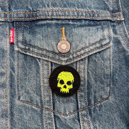 A black felt badge with a yellow embroidered skull attached to the front pocket of a blue denim jacket. The badge is prominently displayed against the denim fabric