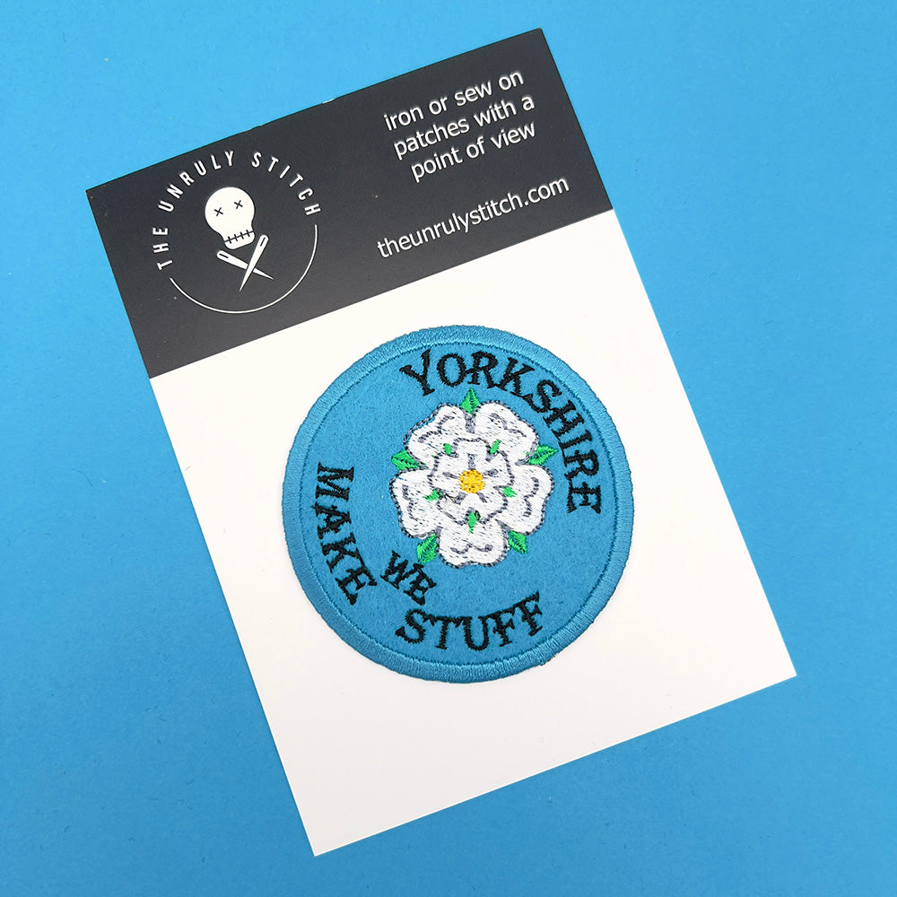 The same patch with a light blue background and white rose, displayed on a white card with "The Unruly Stitch" branding on a black header. The card sits on a blue background.