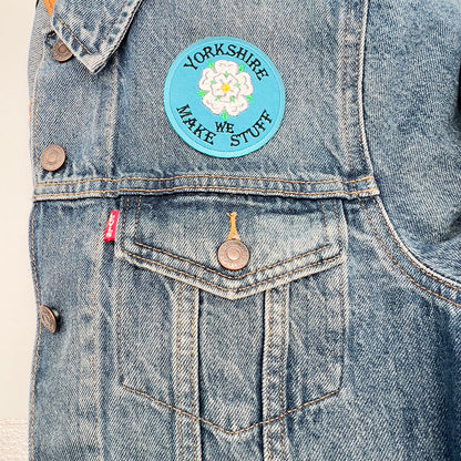 The round patch with a light blue background and white rose is ironed onto a blue denim jacket, positioned above the left breast pocket.