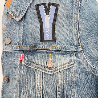  An embroidered felt patch shaped like an uppercase "Y" attached to the front of a denim jacket. The patch features a blue zipper with white teeth and a white pull tab, depicted with black outlines and details. The jacket shows buttons and a visible pocket flap.