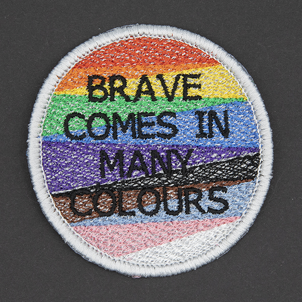Round embroidered patch, embroidered with a silver border and pride rainbow stripes. The words BRAVE COMES IN MANY COLOURS in black text sit in top of the stripes.Photographed on a black background