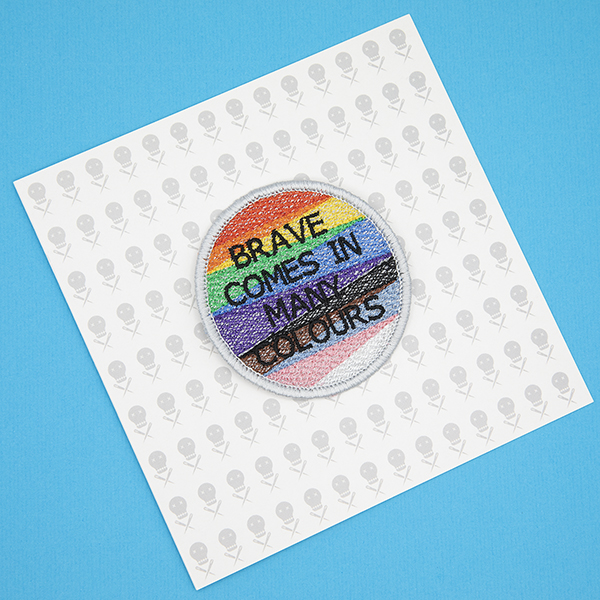 Round embroidered patch, embroidered with a silver border and pride rainbow stripes. The words BRAVE COMES IN MANY COLOURS in black text sit in top of the stripes.Photographed on a gift card printed with tiny images of The Unruly Stitch logo