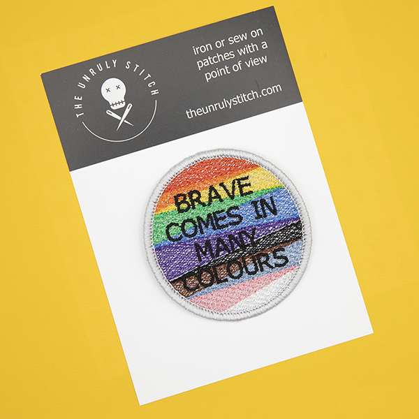 Round embroidered patch, embroidered with a silver border and pride rainbow stripes. The words BRAVE COMES IN MANY COLOURS in black text sit in top of the stripes.Photographed on a black and white postcard with The Unruly Stitch logo