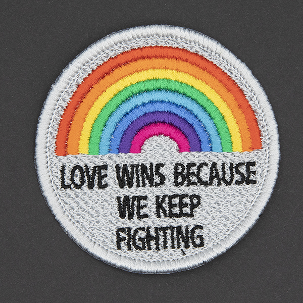 round embroidered patch, embroidered in silver with an eight colour rainbow and the words LOVE WINS BECAUSEWE KEEP FIGHTING in black text photographed on a black background