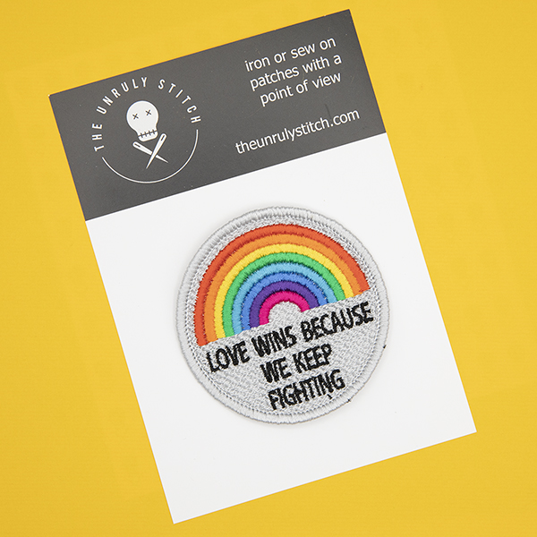 round embroidered patch, embroidered in silver with an eight colour rainbow and the words LOVE WINS BECAUSEWE KEEP FIGHTING in black text photographed ona black and white postcard with The Unruly Stitch logo