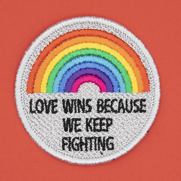 round embroidered patch, embroidered in silver with an eight colour rainbow and the words LOVE WINS BECAUSEWE KEEP FIGHTING in black text photographed on a red background