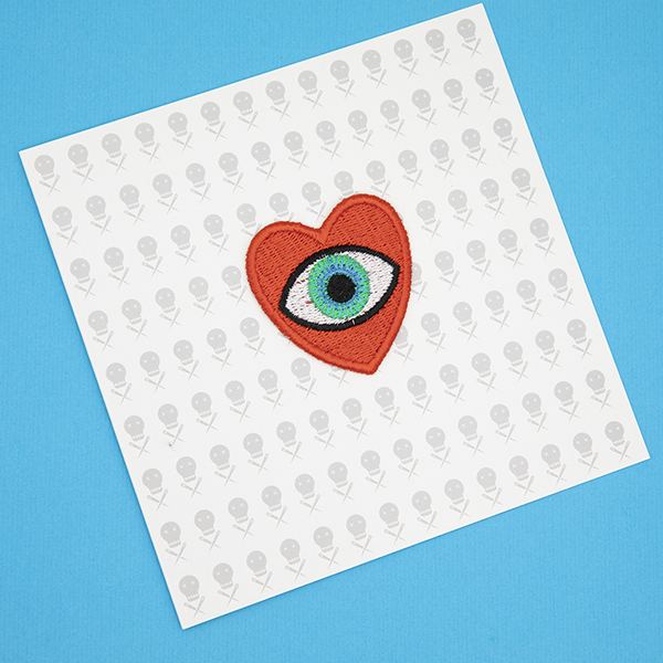 medium embroidered patch, red heart containing a blue eye photographed on a gift card printed with tiny images of The Unruly Stitch logo