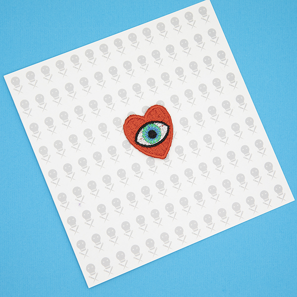 small embroidered patch, red heart containing a blue eye photographed on a gift card with tiny images of The Unruly Stitch logo