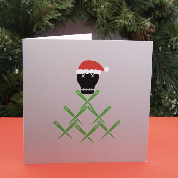 Front of card for decorations - the Unruly Stitch skull wears a santa hat and sits on a pile of green sewing needles, shown standing on a red surface with a fir wreath in the background