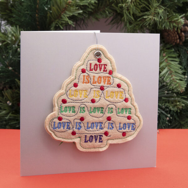 cream felt ornament, Christmas tree shape embroidered with LOVE IS LOVE in pride rainbow colours and red fairy lights , shown standing on a red surface with a fir wreath in the background