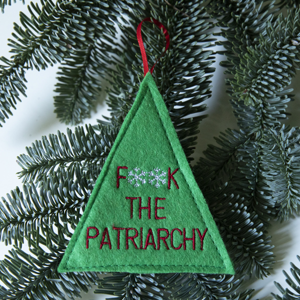 green felt Christmas tree shape embroideredwith the words F**K THE PATRIARCHY. ** are snowflakes. Thedecoration is finished with a red ribbon and is shown against a pine branch.
