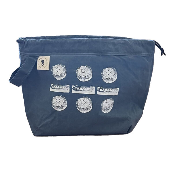 large slate blue drawstring project bag. The bag is printed with Tunnocks tea cakes and caramel wafers spelling out the dots and dashes for SOS in white ink. The bag has a handle, a drawstring and a cream label with The Unruly Stitch logo