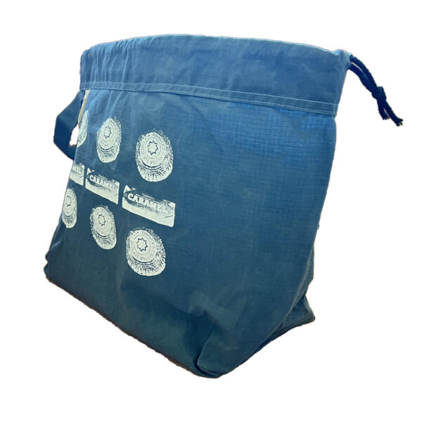 side view of large slate blue drawstring project bag. The bag is printed with Tunnocks tea cakes and caramel wafers spelling out the dots and dashes for SOS in white ink. The bag has a handle, a drawstring and a cream label with The Unruly Stitch logo